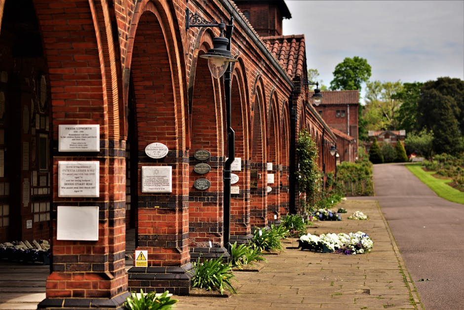 A row of red-brick buildings with arched doorways and black wrought iron lanterns mounted on the walls, situated alongside a paved walkway in a peaceful garden setting. In front of the buildings, ther