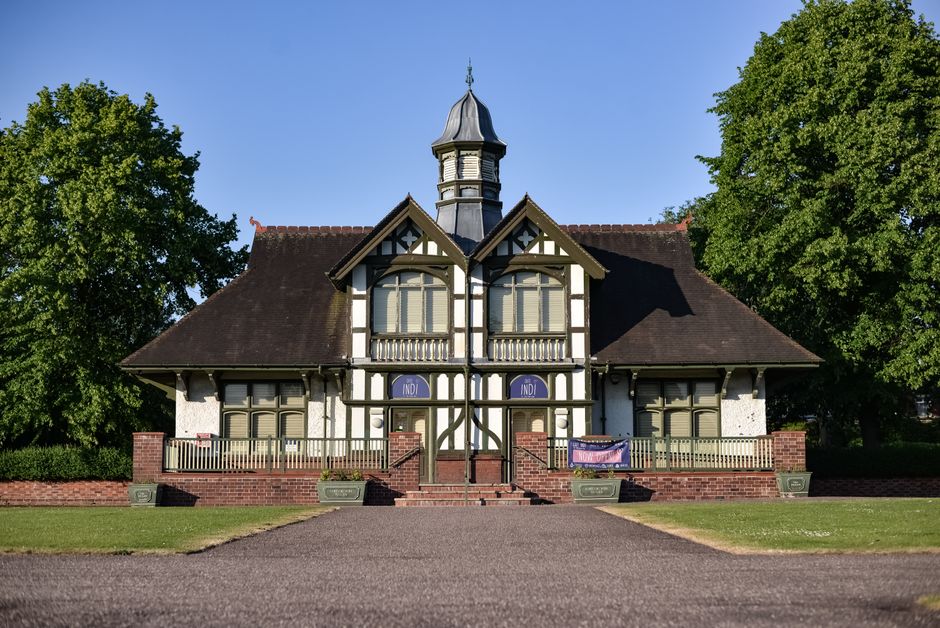 A row of red-brick buildings with arched doorways and black wrought iron lanterns mounted on the walls, situated alongside a paved walkway in a peaceful garden setting. In front of the buildings, ther