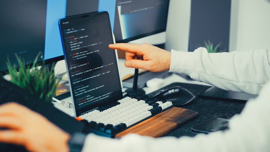 A close-up view of a person's hand pointing at a code editor displayed on a smartphone screen, which is set up on a wooden desk alongside a mechanical keyboard with white and black keys, a wireless mo
